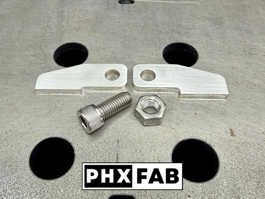 Two stainless steel tabs with pre-drilled holes, a matching stainless threaded bolt, and a hex nut, arranged on a gray metal surface with black circular cutouts. A PHX FAB logo overlay is at the bottom.